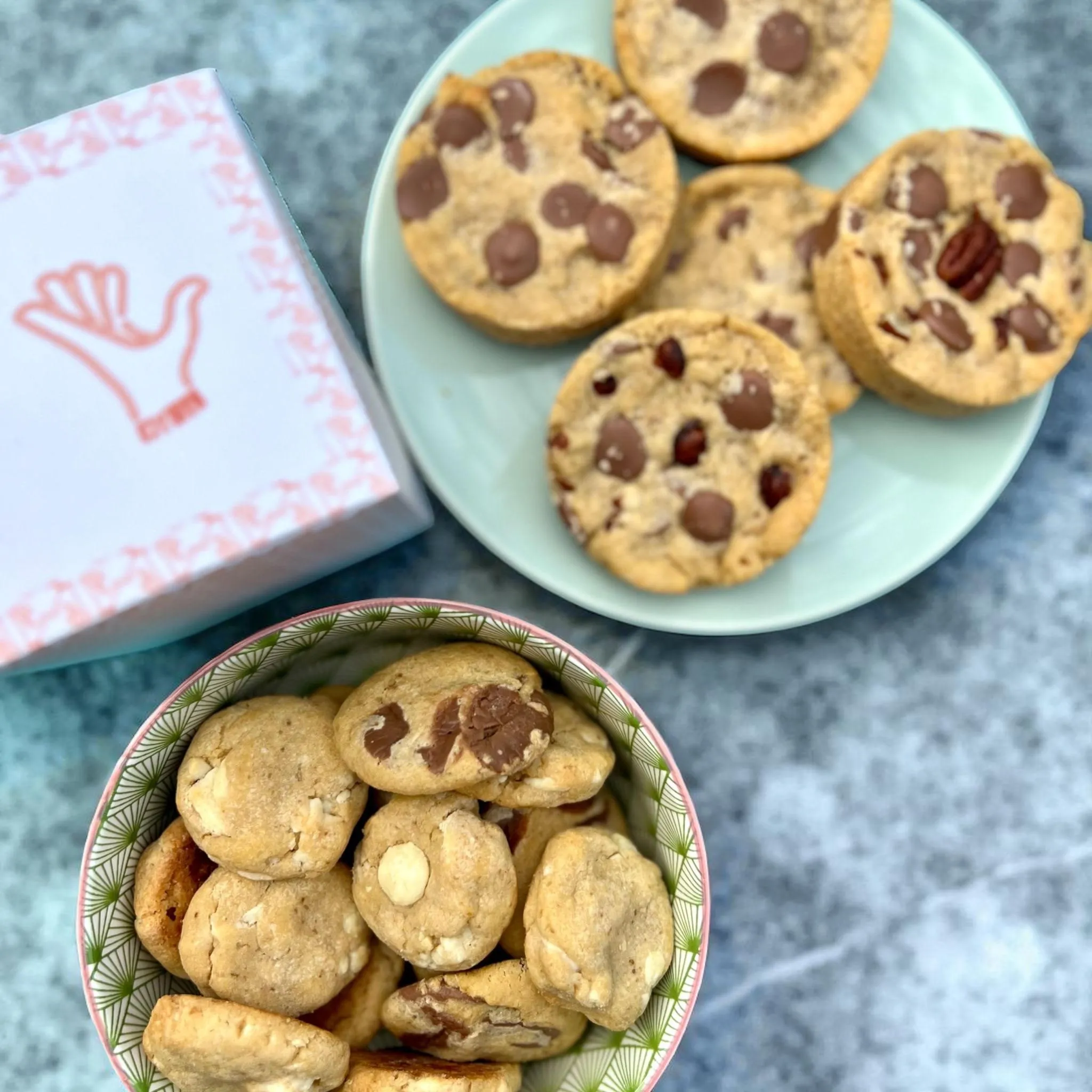 Cookies aux pépites de chocolat et aux noix de pécan sur une assiette, coupelle de mini-cookies et boîte Mani & Co posées sur un plan de travail